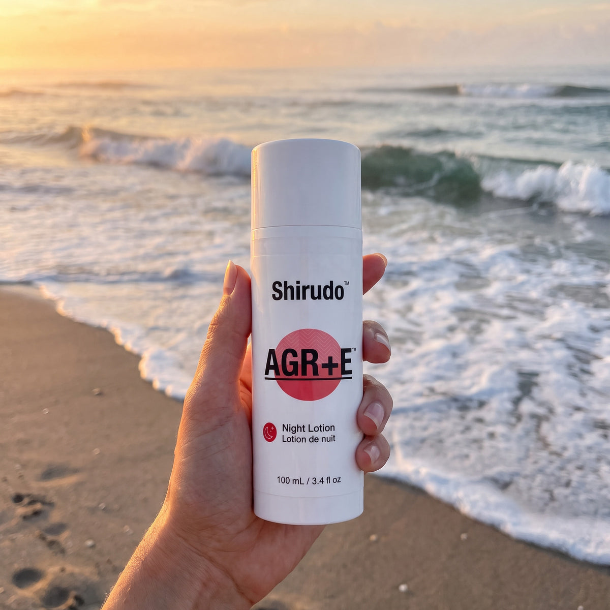 A hand holds a bottle of Shirudo AGR+E Night Lotion against a blurred beach background at sunset.